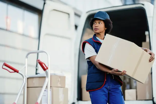 Black female courier unloading cardboard boxes from a van while making a delivery in the city.